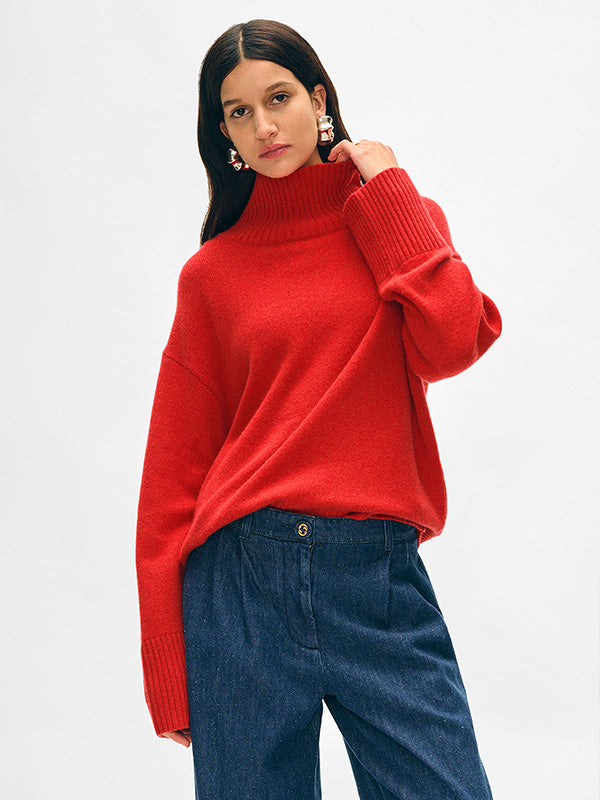 Model wears the White + Warren Cashmere Easy Standneck sweater in Carmine Red with pleated pants and red sneakers while standing in front of a stone wall.